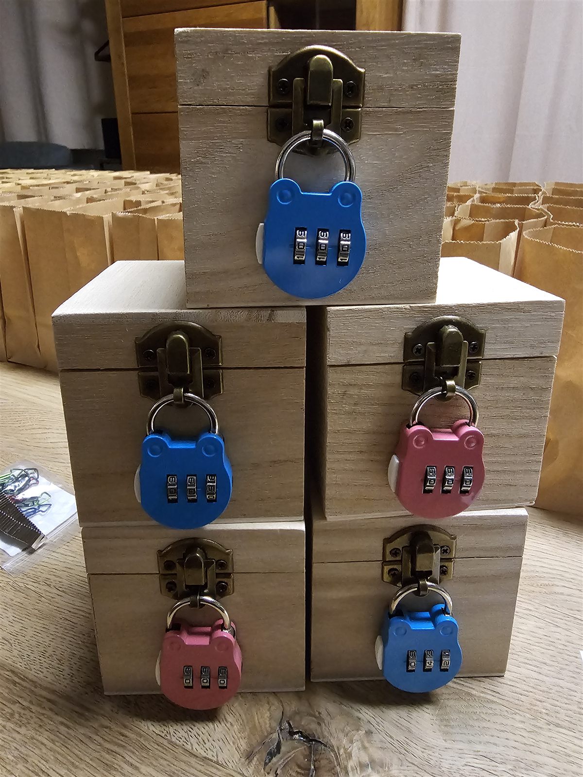 Fünf kleine Holzboxen mit blauen und rosa dreistelligen Zahlenschlössern vor braunen Papiertüten.
