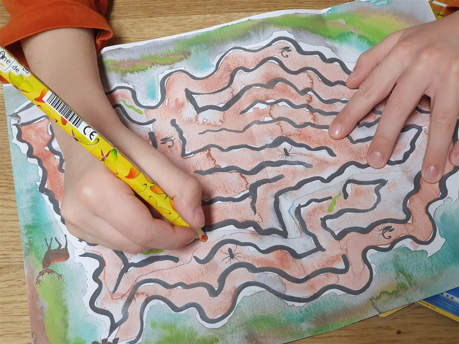 Ein Kind zeichnet mit einem gelben Stift den Weg durch ein buntes Labyrinth auf Papier.