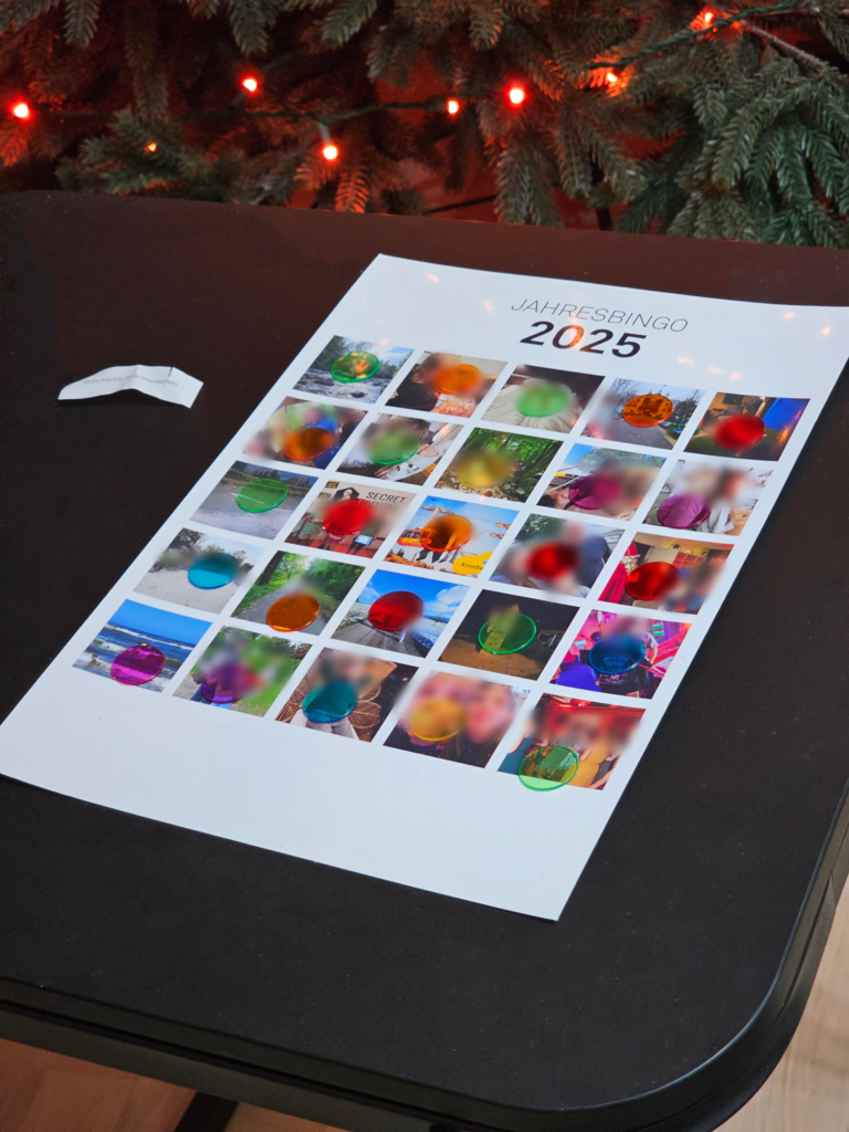 Jahresbingo 2025 mit eigenen Fotos und bunten Spielplättchen auf einem Tisch vor einem beleuchteten Weihnachtsbaum.